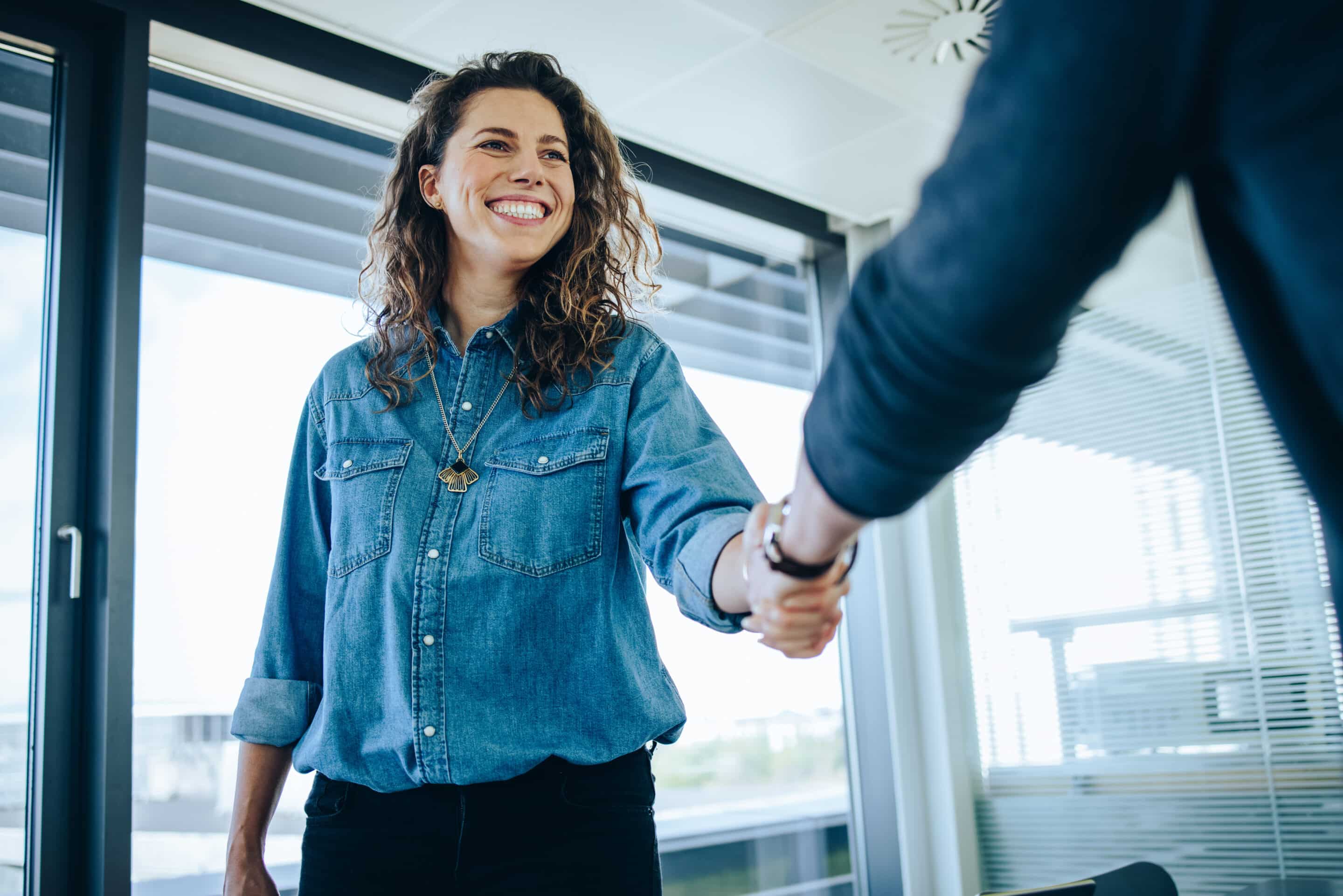 Femme de face souriante qui est entrain de faire une poignée de main à une autre personne vue de dos.
