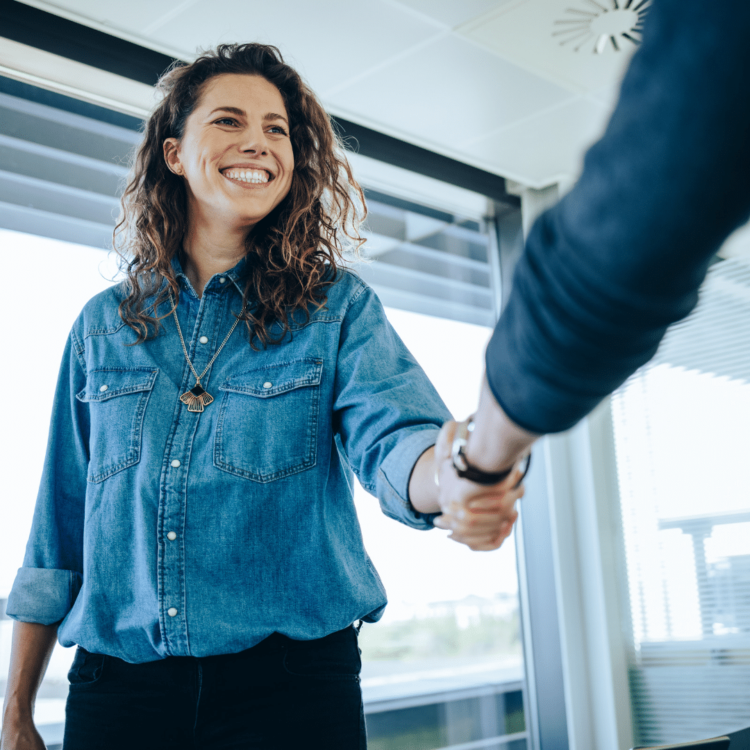 Femme de face souriante qui est entrain de faire une poignée de main à une autre personne vue de dos.